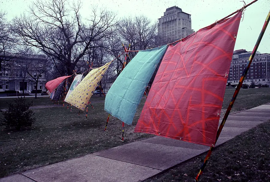 Painted fabric panels in red, aqua and yellow hung from striped poles on a lawn with bare trees and city buildings behind.