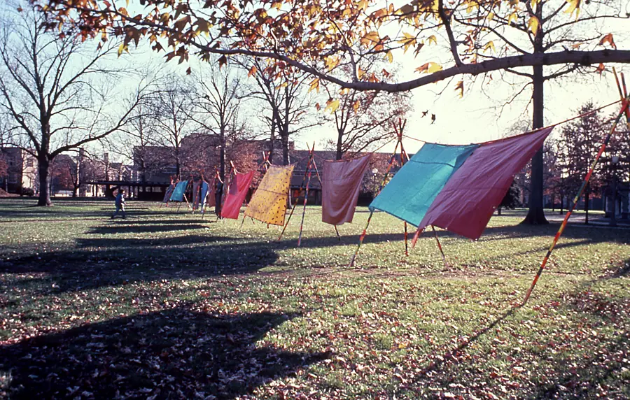 Row of painted fabric panels on striped poles across an autumn lawn under a tree with orange leaves.