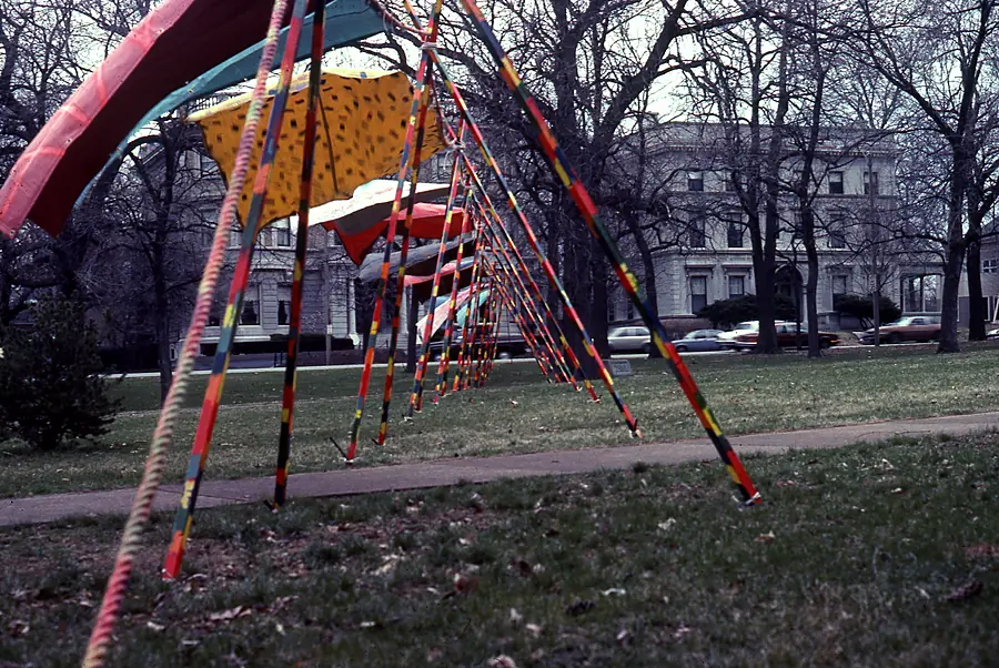 Line of striped poles carrying painted fabric panels across a park lawn with bare trees and a large stone house behind.