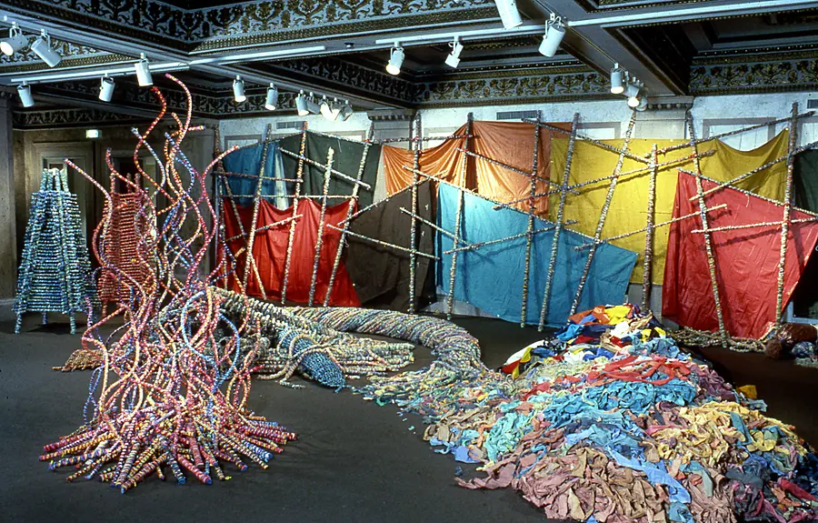 Panels of colored fabric on a pole lattice, a fountain of curved rods and a long coil of wrapped rope across a dark floor.