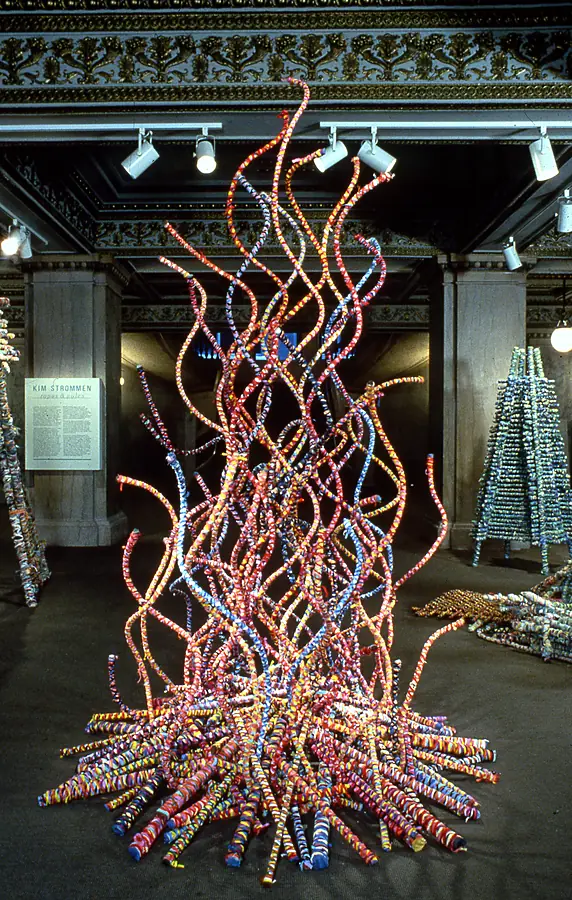 Tall flame-shaped cluster of curved wrapped rods rising from a pile of horizontal poles, beside a wall label reading Kim Strommen, ropes and poles.