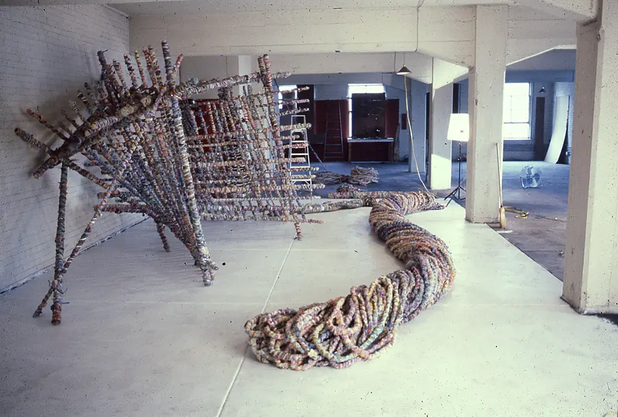 Long wrapped-rope form curving across a concrete floor beside a leaning lattice of wrapped poles.