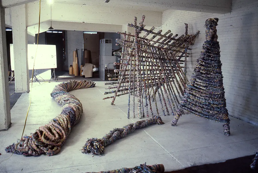 Coiled rope on the floor, a slanted lattice of wrapped poles and a tall conical form in a white brick room.