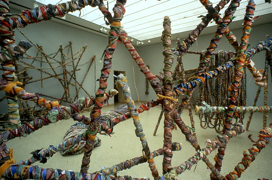 View through foreground poles wrapped in red, orange, blue and green strips into a skylit gallery of more wrapped structures.