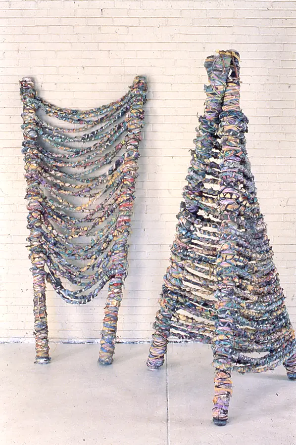 Two forms of wrapped strips, one hung like a curtain of loops and one an A-frame cone, in front of a white brick wall.