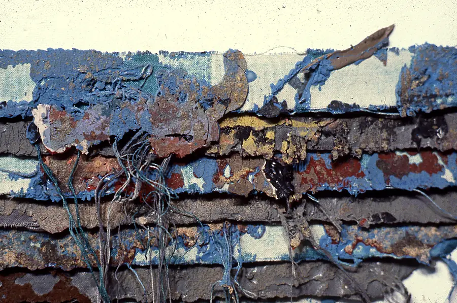 Detail of the edge of a layered work: paint-heavy canvas strips in blue, rust, mustard and gray with loose threads.