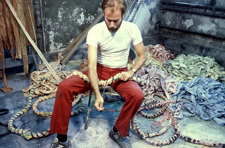Bearded man in a white T-shirt and red trousers seated in a studio, winding a thick rope with strips of dyed cloth.