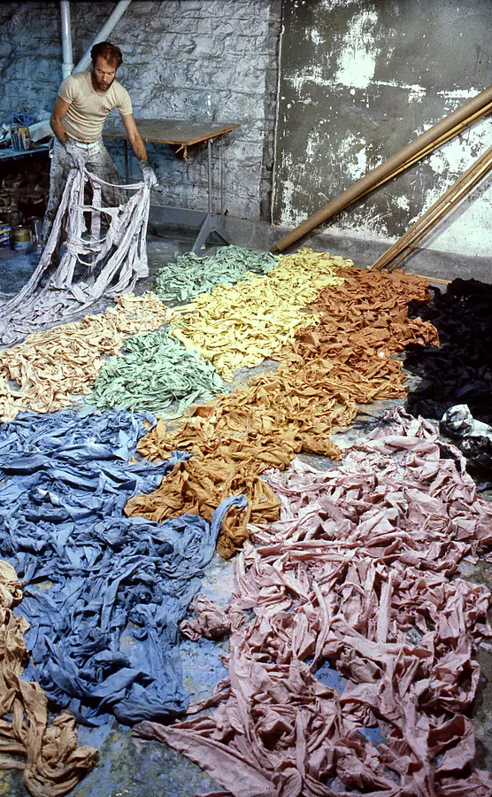Man in a basement studio lifting a bundle of lilac fabric strips above floor heaps of dyed strips sorted by color.