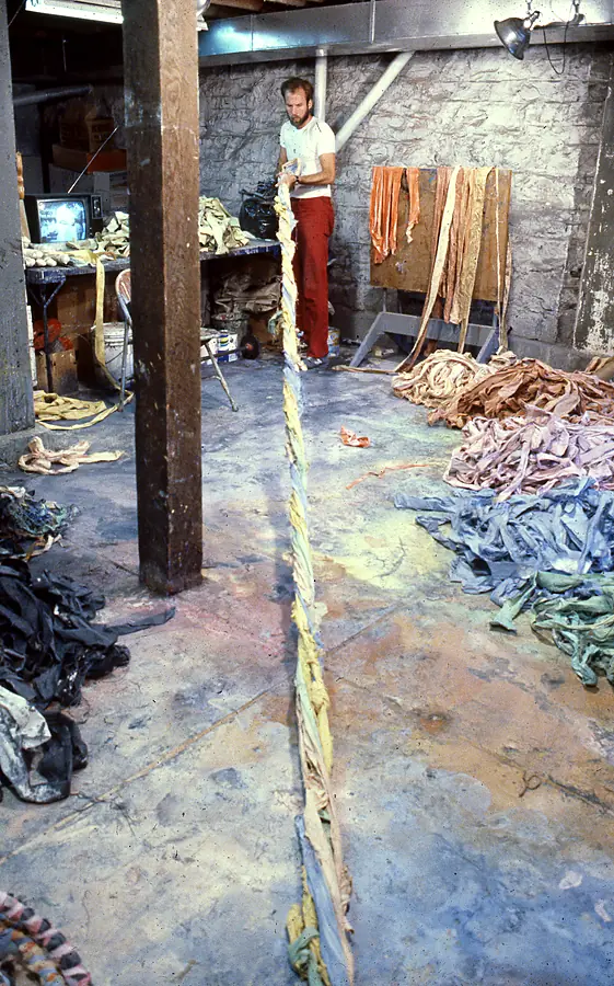Man standing at the far end of a very long twisted rope of cloth strips laid across a paint-stained studio floor.