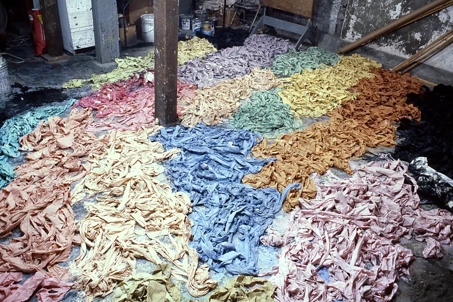 Studio floor covered in heaps of dyed fabric strips arranged in patches of pink, blue, tan and black.