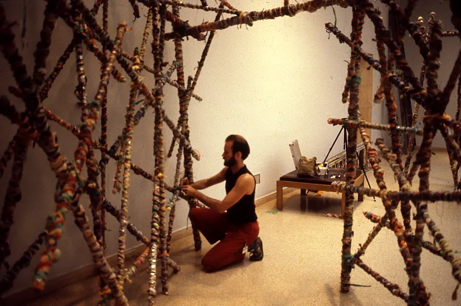 Man kneeling inside a lattice of wrapped poles in a gallery, working on the structure.