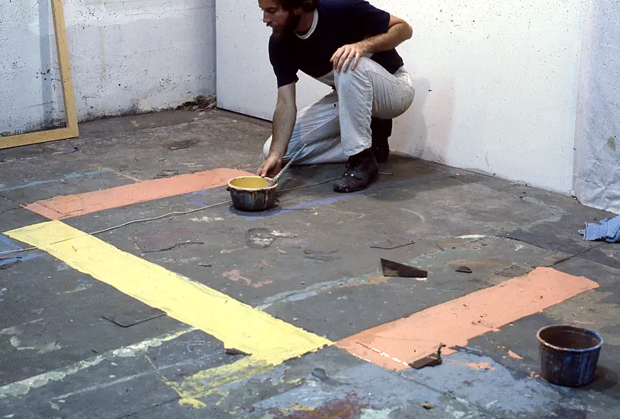 Bearded man crouching in a studio beside a can of yellow paint and broad bands of yellow and salmon paint on the floor.