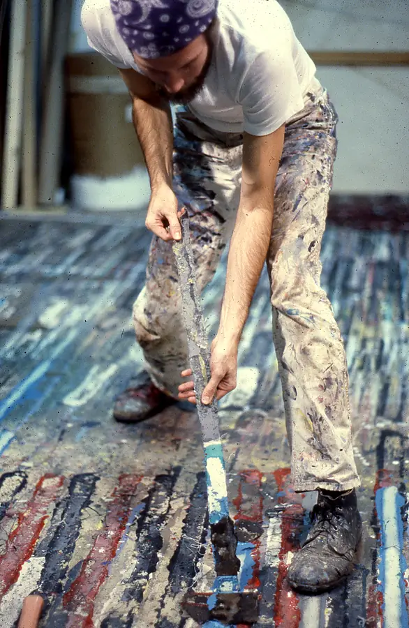 Bearded man in a purple bandana lifting a long strip of dried paint from a striped studio floor.