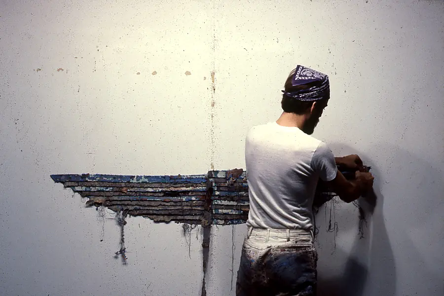 Man in a bandana at a white wall, attaching a horizontal strip of layered blue and brown paint.