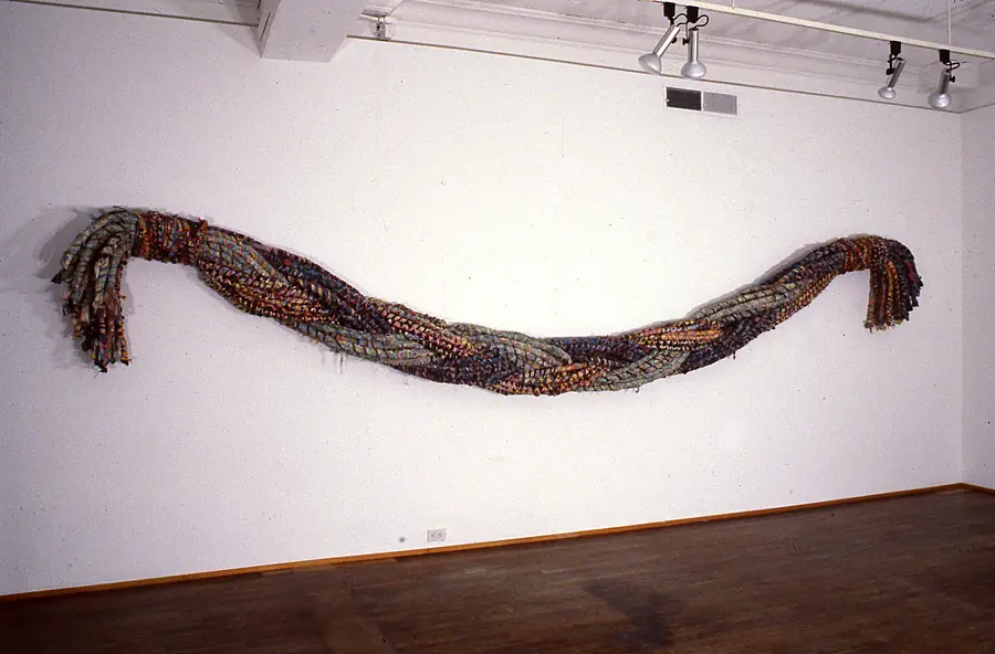 Long twisted braid of painted canvas strips hung across a gallery wall, its ends fraying into bundles.