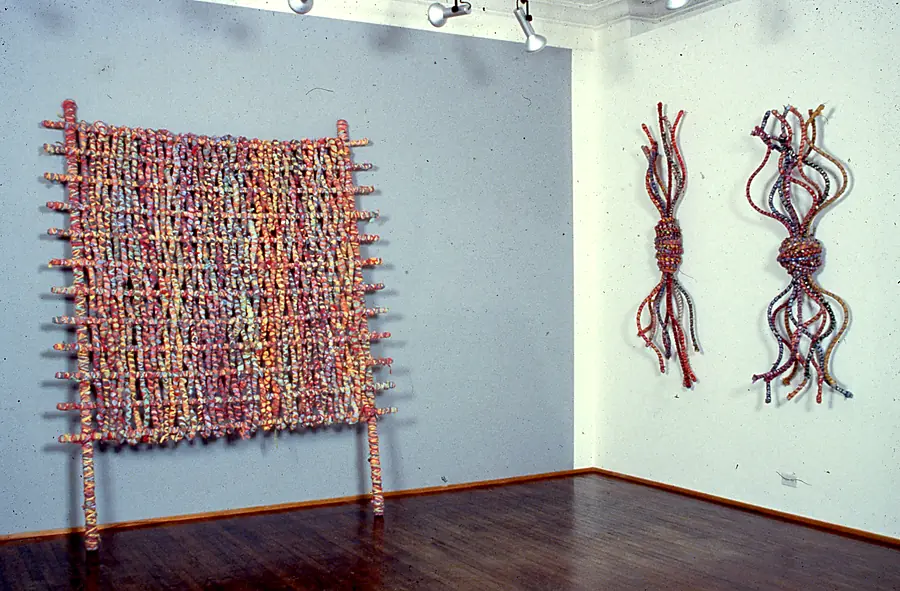 Gallery corner with a woven grid of wrapped poles on legs and two wall-hung bundles with knotted waists and trailing ends.