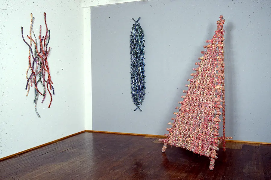 Gallery corner with wavy wrapped cords on the wall, a blue woven spindle and a pink triangular woven frame on short legs.
