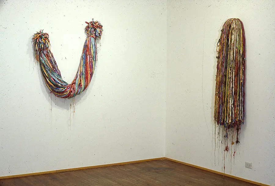 Two wall pieces in a gallery corner: a U-shaped bundle of colored strips and a long curtain of hanging strands.