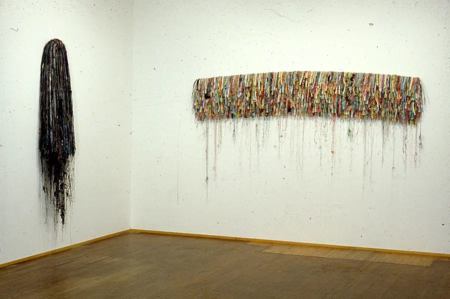 Two wall-mounted works of hanging fabric strips, one tall and dark, one a long multicolored band with threads trailing below.