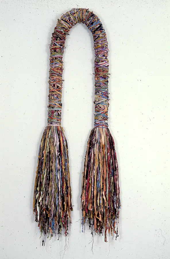 Tall wall-hung arch wrapped in multicolored strips that ends in two long tassels of shredded cloth.