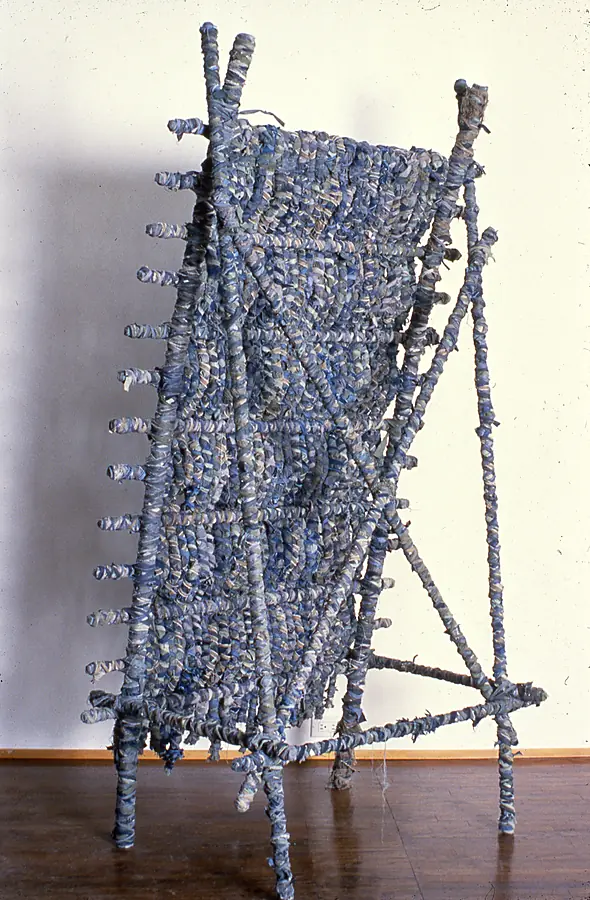 Freestanding woven panel of blue cloth strips on a frame of wrapped poles, leaning against a wall.