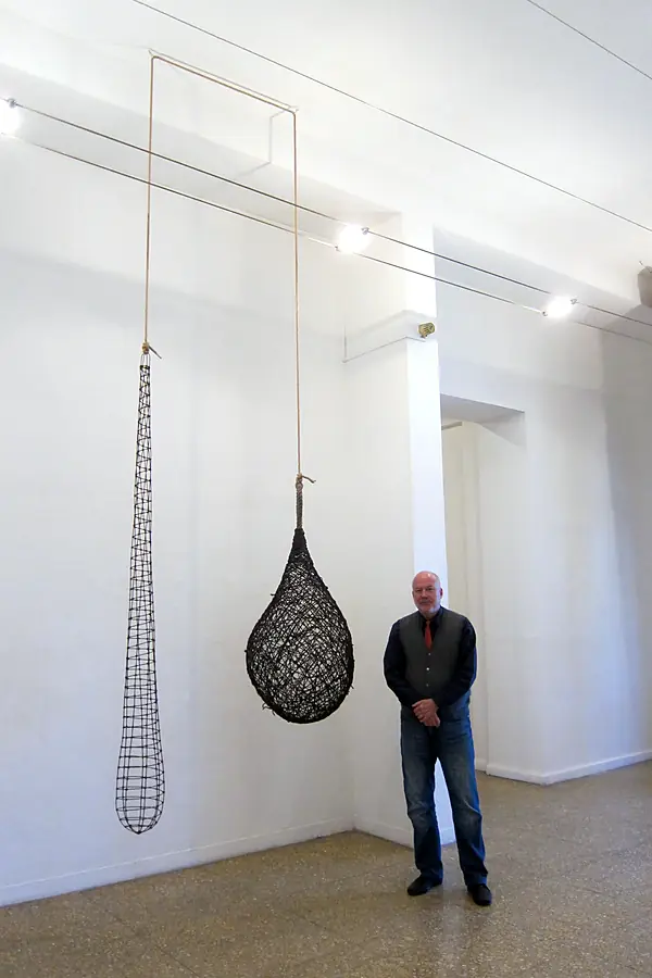 Kim Strommen standing beside two hanging black woven forms in a tall white gallery.