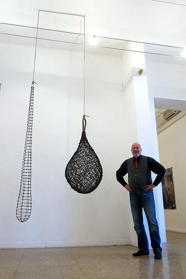 Kim Strommen, smiling, standing with his hands on his hips beside two hanging black woven forms in a white gallery.