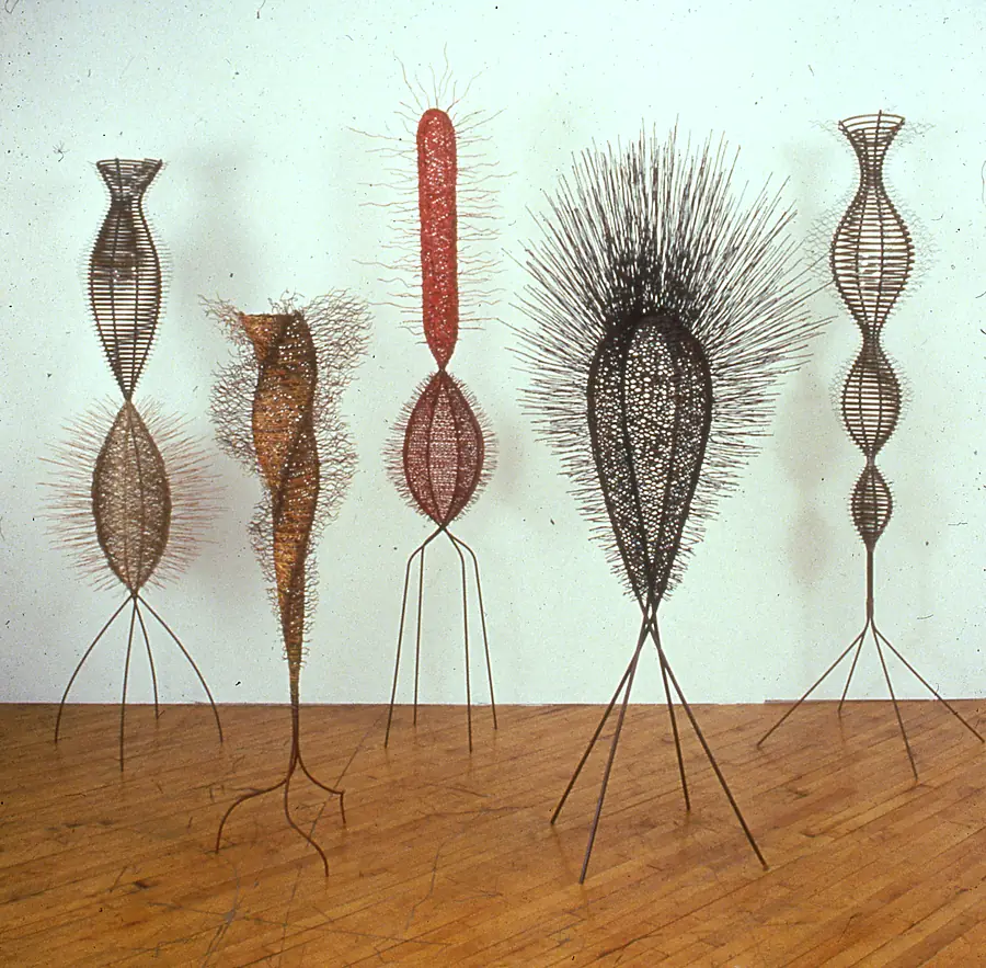 Five tall wire and metal-band figures with spiked halos standing on slender legs in a row against a cream wall.