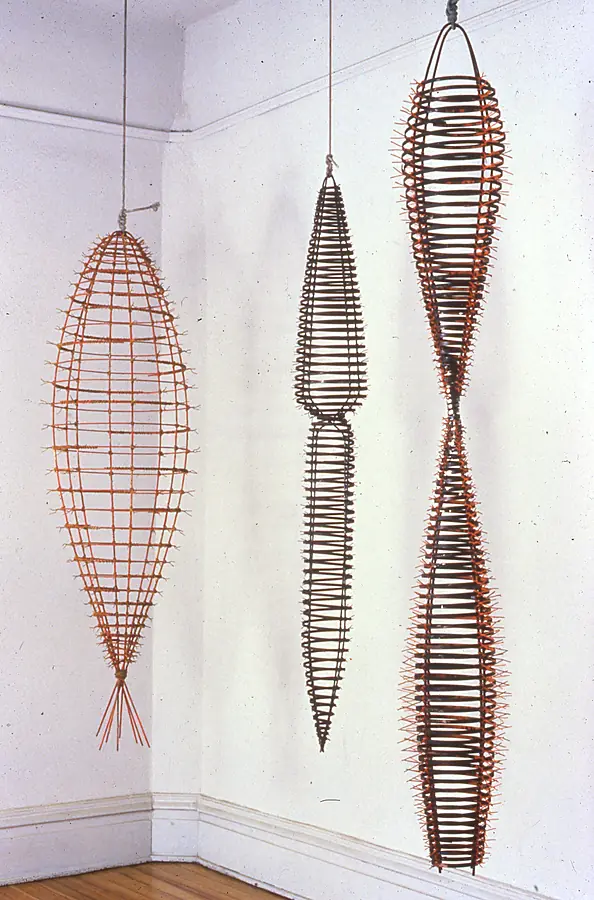 Three hanging ribbed cage forms in orange and dark metal suspended in a white room corner.