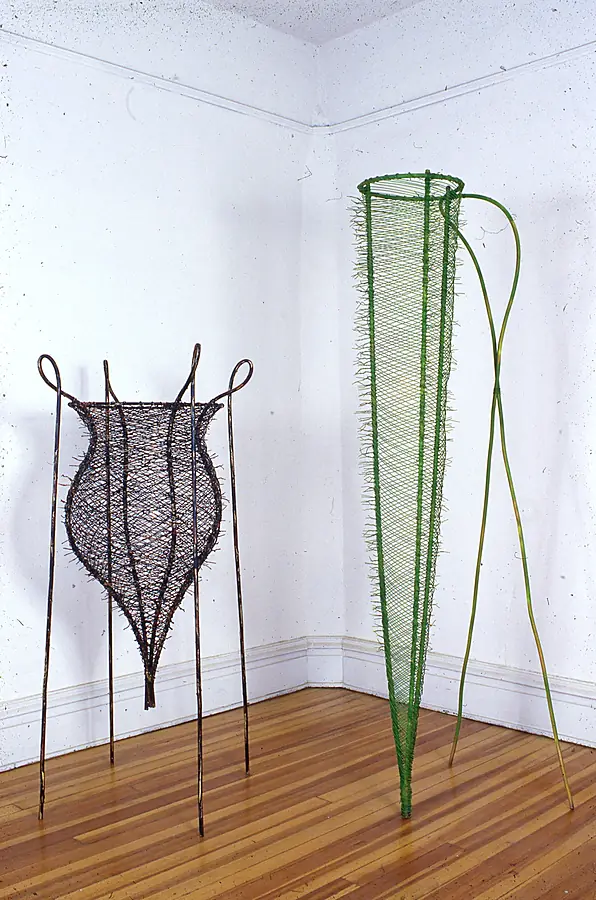 Dark woven wire urn on four legs beside a tall green wire cone in a white room corner.