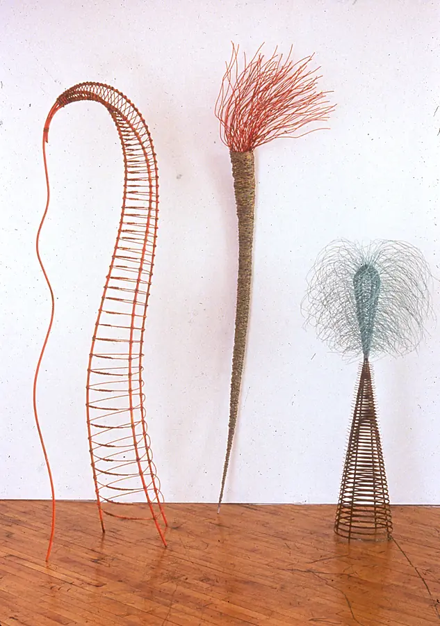 Three tall standing forms in orange, olive and blue-green wire, topped with brush-like tufts.