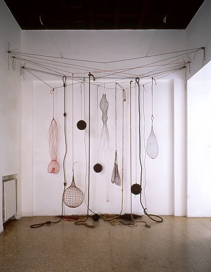 Hanging wire baskets, spheres and nets strung from lines across a white gallery corner, with ropes coiled on the floor.