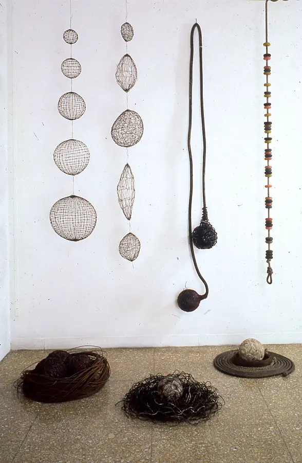 Strings of wire-mesh spheres and a rope of colored discs hanging above floor nests of reed, wire and rope.