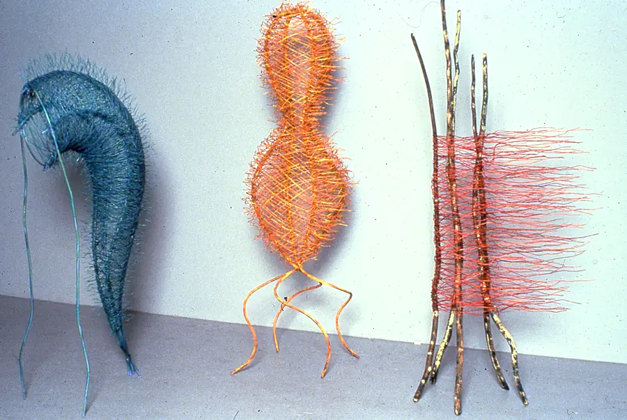 Three standing forms: a blue-green wire crescent, an orange wire gourd on curved legs, and branches woven with red wire.