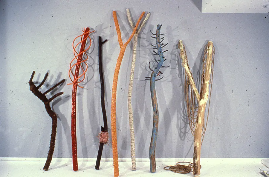 Seven tall branches wrapped in orange, red, cream and brown cord, leaning against a pale wall.