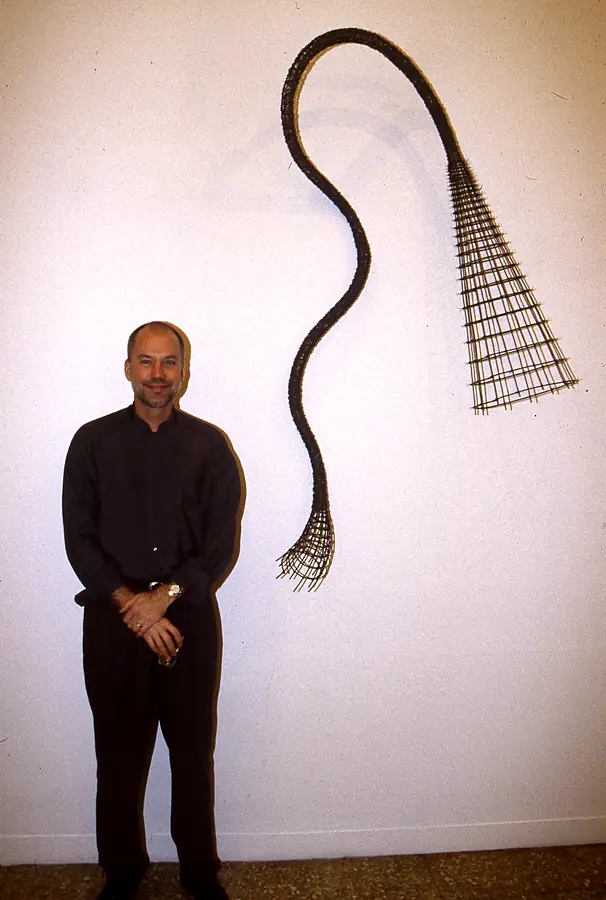 Kim Strommen standing beside a long curving wall-hung form of dark woven mesh that fans into a grid at one end.
