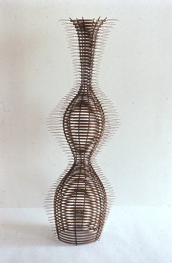 Tall vase-shaped sculpture of stacked copper and rust metal bands with fine wire spikes, against a cream wall.